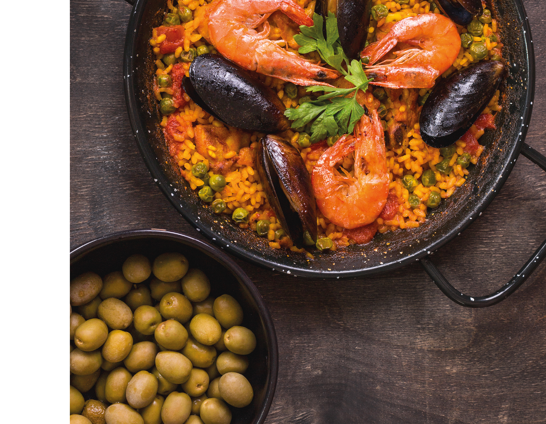 Paella in black pan with rice, shrimps, mussels, squid and meat, bowl with olives and vintage cutlery. Seafood paella, traditional spanish dish. Paella on rustic table. Selective focus. Top view
