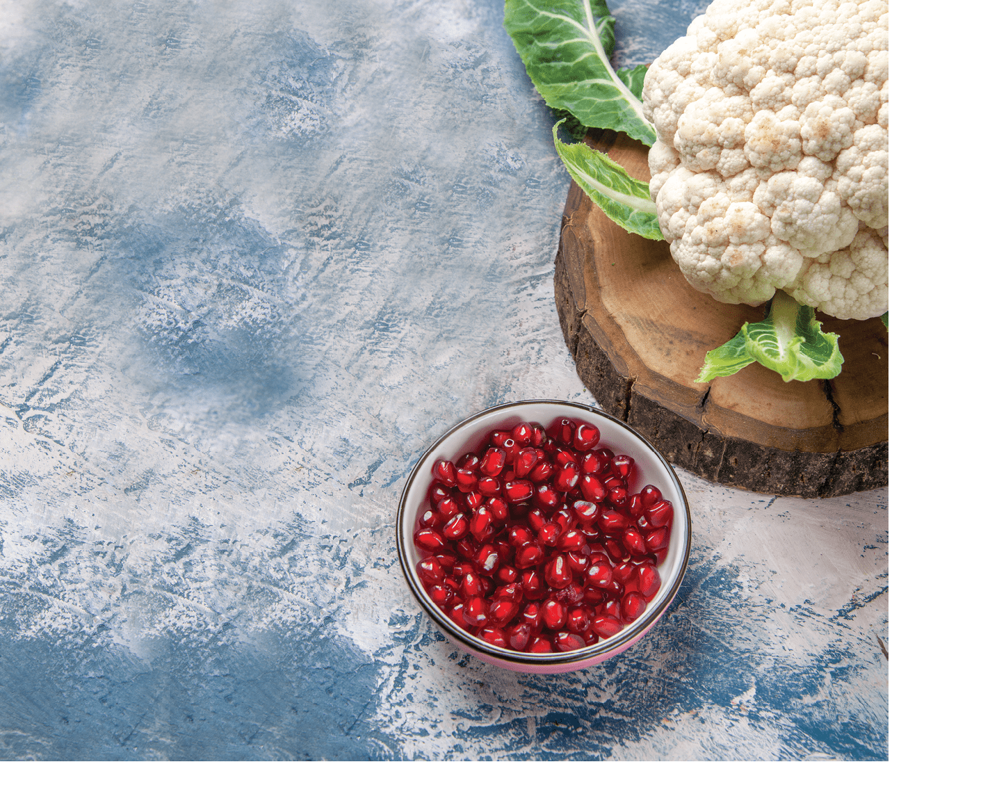 Vertical view of fresh cauliflower with leaves on brown wooden tray broccoli and pomegranate on gray background