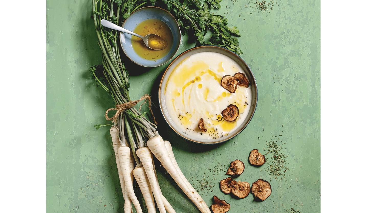 Parsnip cream soup in ceramic bowl with sun dried pears, butter sauce, bundle of fresh parsnip and herbs over green texture background. Flat lay, space