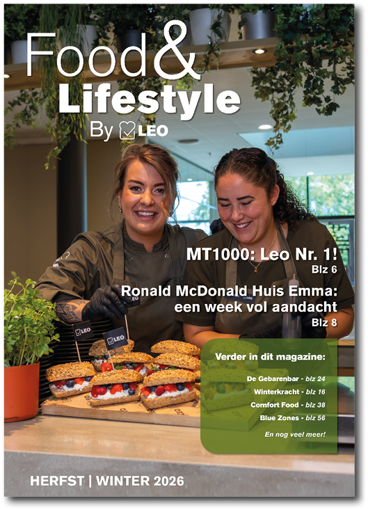 Two adults wearing dark chef jackets smile and prepare sandwiches with fresh ingredients in a bright kitchen setting for a food and lifestyle magazine cover. Description generated by AI