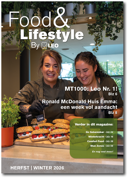 Two adults wearing dark chef jackets smile and prepare sandwiches with fresh ingredients in a bright kitchen setting for a food and lifestyle magazine cover. Description generated by AI