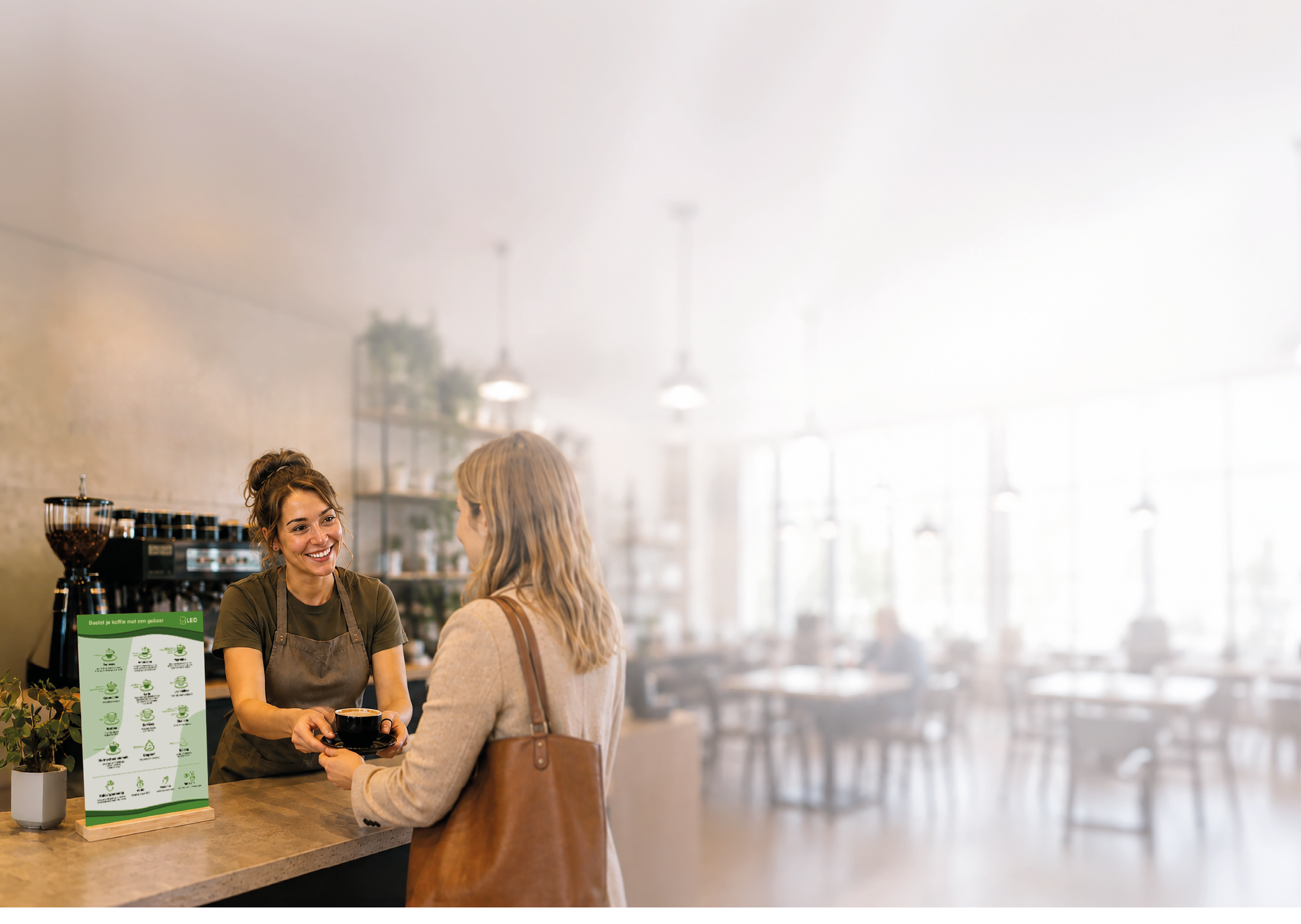 An adult with a ponytail and apron is smiling and handing a cup to another adult with long hair and a shoulder bag across a coffee shop counter. Description generated by AI