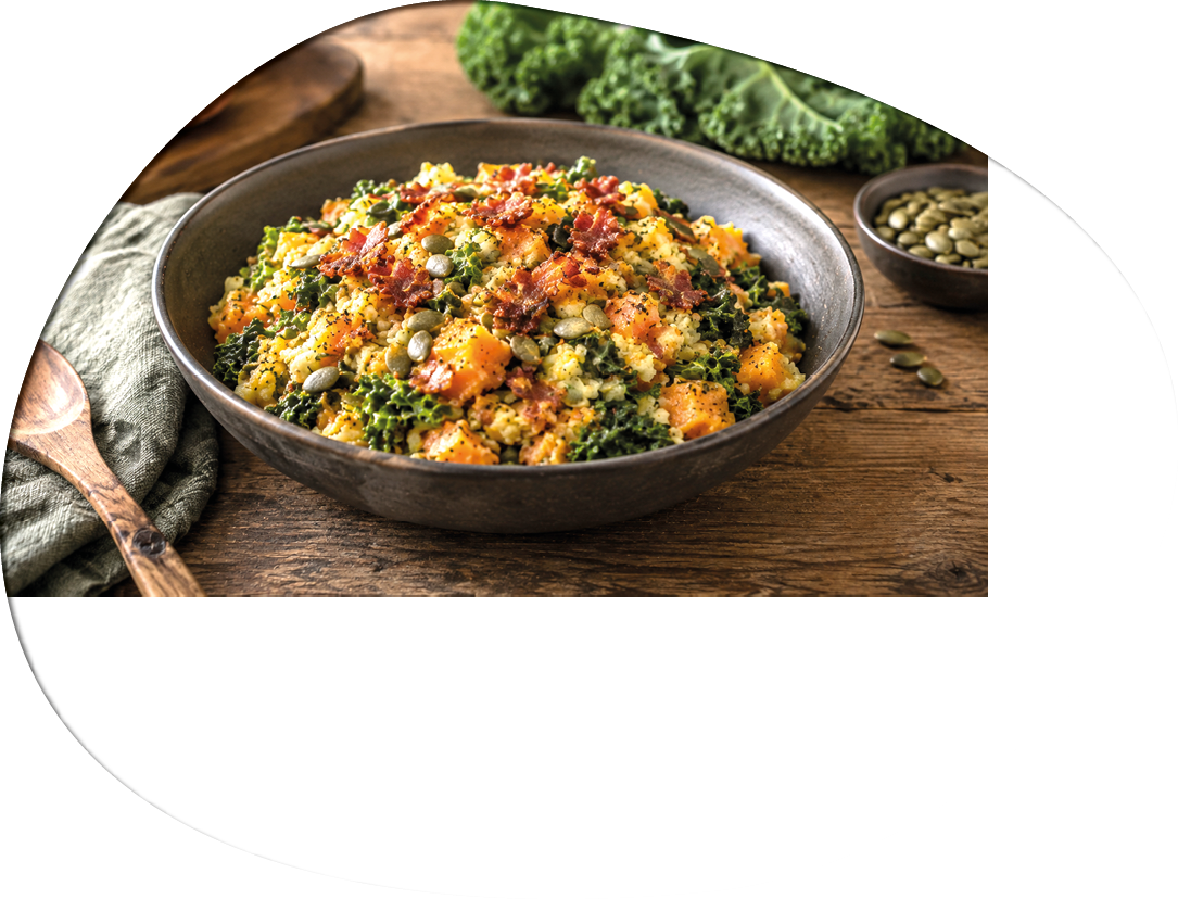 A bowl of cooked quinoa mixed with kale, roasted butternut squash, pumpkin seeds, and crispy bacon bits on a wooden table with a wooden spoon and a small bowl of pumpkin seeds nearby. Description generated by AI