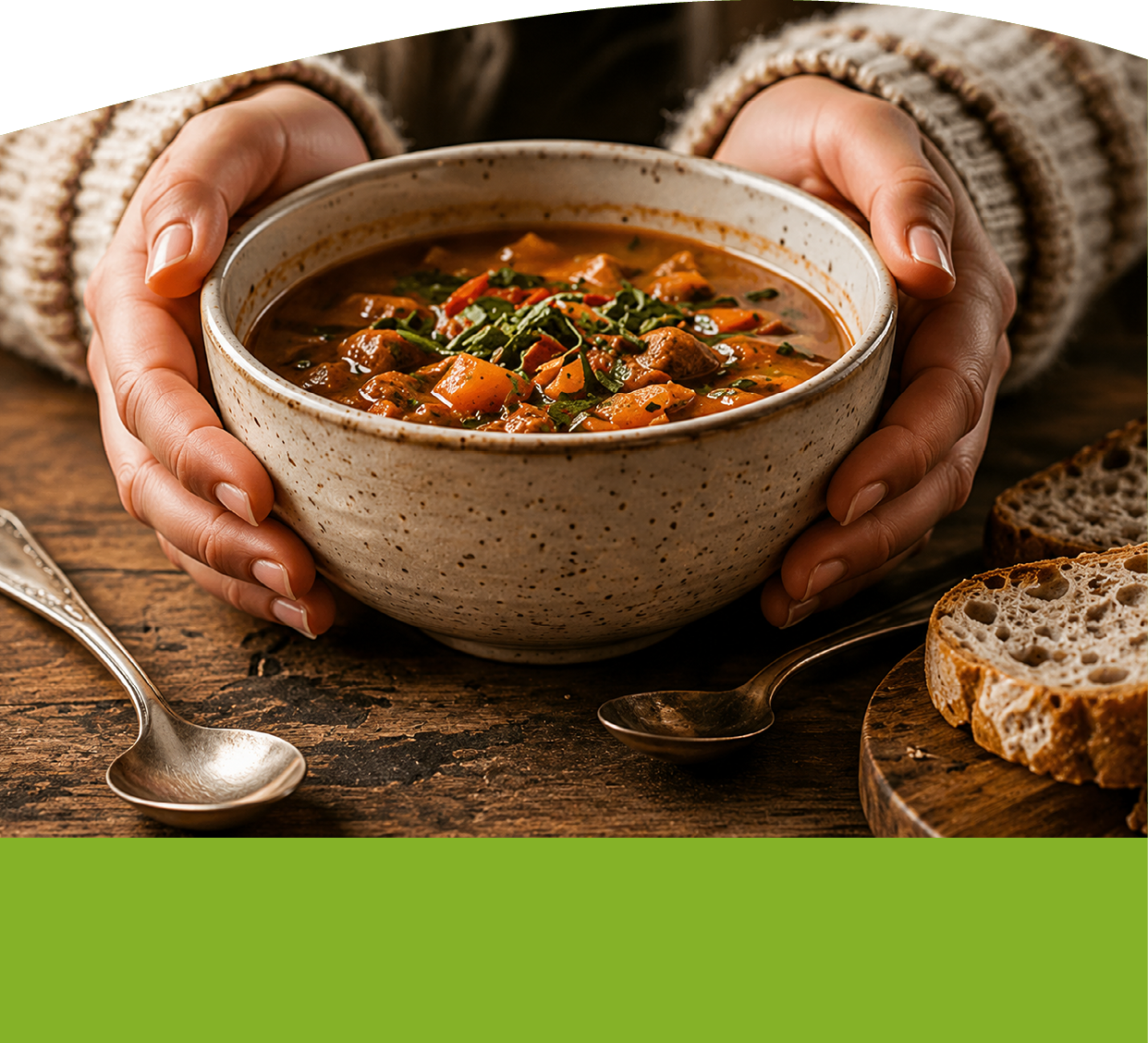 Hands in a cozy sweater are holding a bowl of hearty vegetable and meat soup on a wooden table next to two spoons and slices of rustic bread. Description generated by AI