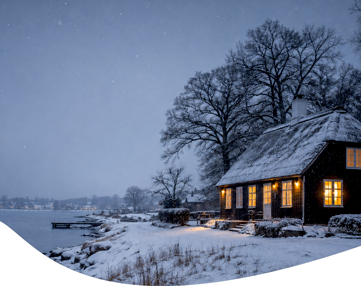 A cozy cabin with glowing windows sits surrounded by snow-covered ground and bare trees near a calm body of water under a gray winter sky. Description generated by AI