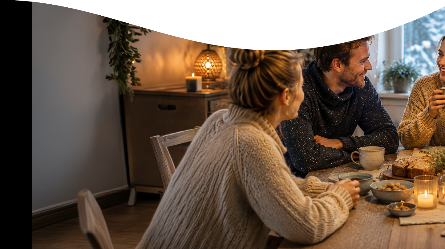 Three people wearing cozy sweaters are sitting around a wooden table with candles, snacks, and drinks, engaged in conversation in a warmly lit room. Description generated by AI