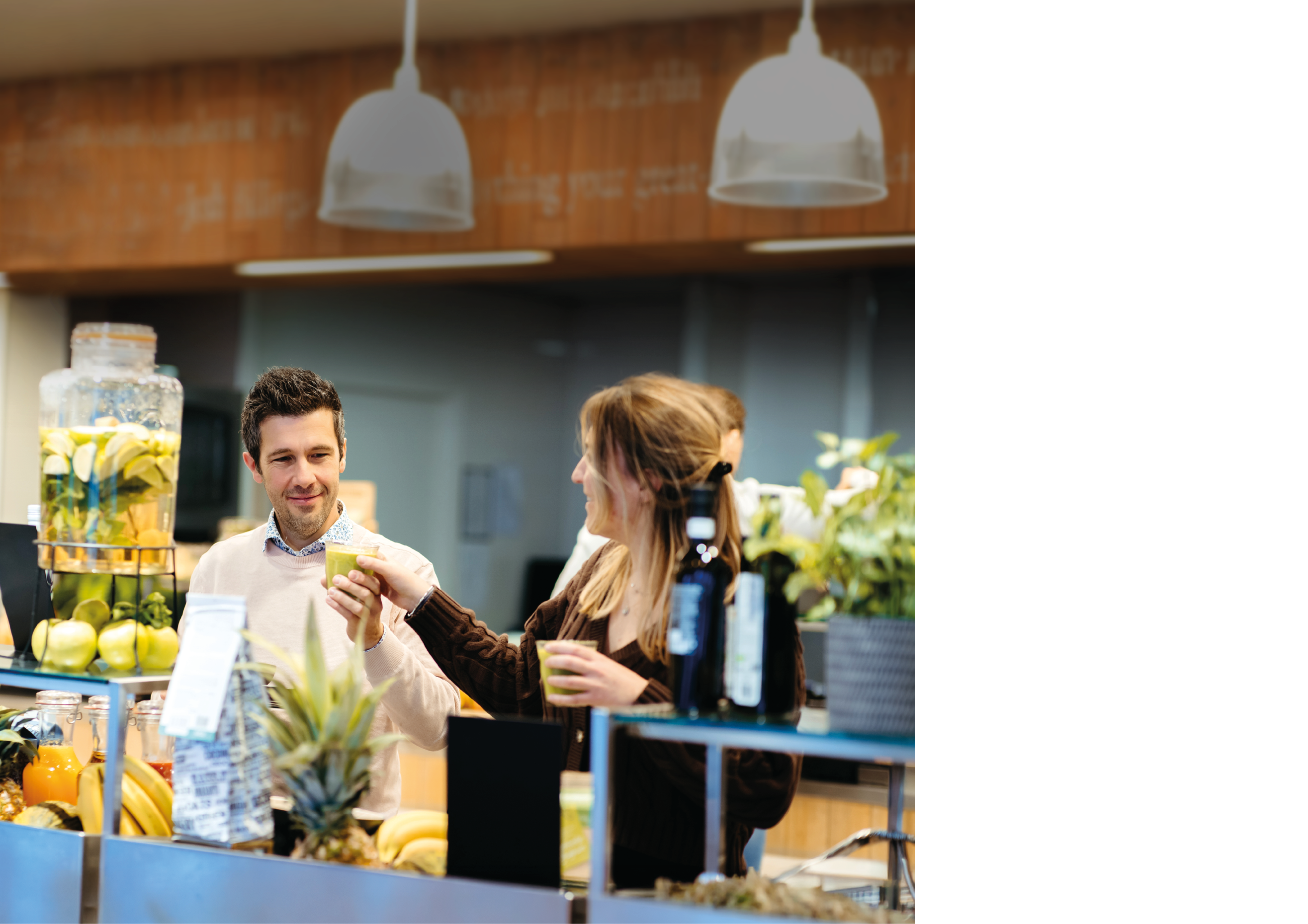 Two adults are smiling and holding drinks while standing near a counter with fresh fruit and beverage containers in a bright indoor setting. Description generated by AI