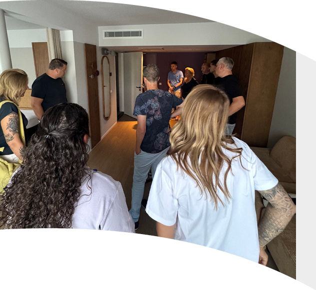 A group of people with varied hairstyles and casual clothing are gathered in a room, some facing a doorway where a few others stand, engaged in conversation. Description generated by AI