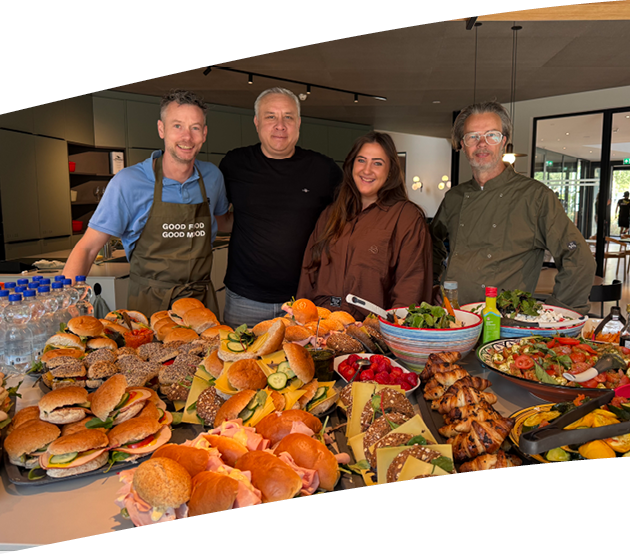 Four adults stand behind a table filled with a variety of sandwiches, salads, grilled chicken, and bottled water in a modern kitchen setting. Description generated by AI