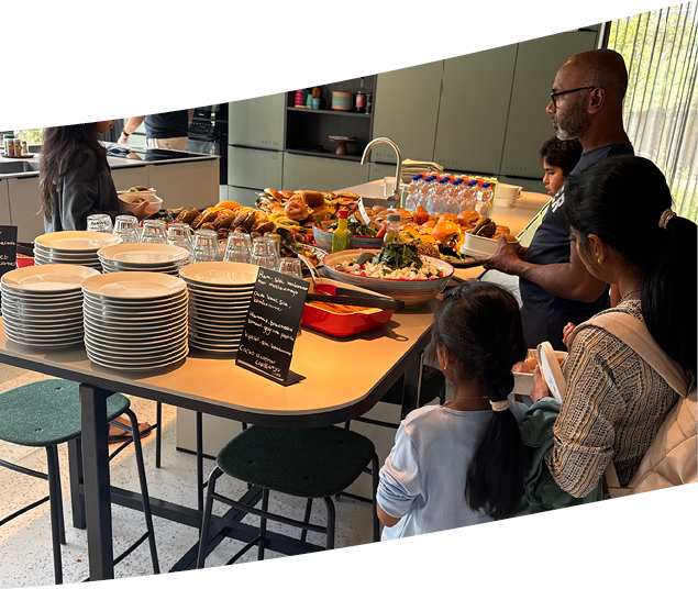 People are gathered around a kitchen island with stacks of plates, glasses, and a variety of food items including sandwiches and fruit, while some hold plates and serve themselves. Description generated by AI