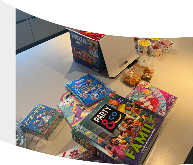 A table displays various colorful board games and snacks including a Mega Box, Party & Co Family edition, and other games alongside cups of snacks and candy. Description generated by AI