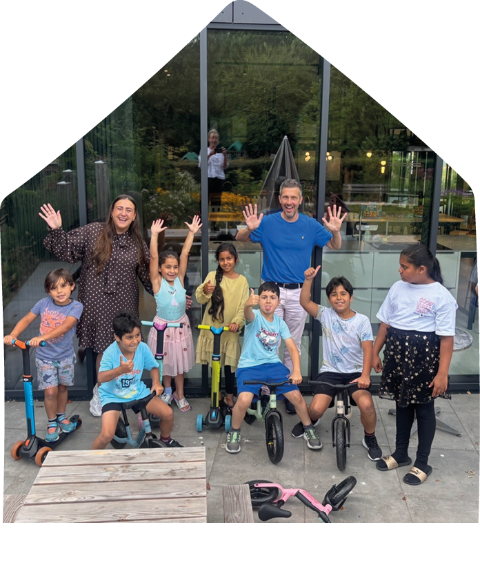 A group of smiling people including children and adults are posing outdoors with some children sitting on scooters and small bikes in front of a glass building. Description generated by AI