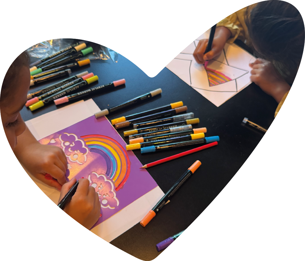 Two children are sitting at a table coloring with markers, one working on a rainbow and cloud drawing and the other filling in a geometric pattern. Description generated by AI