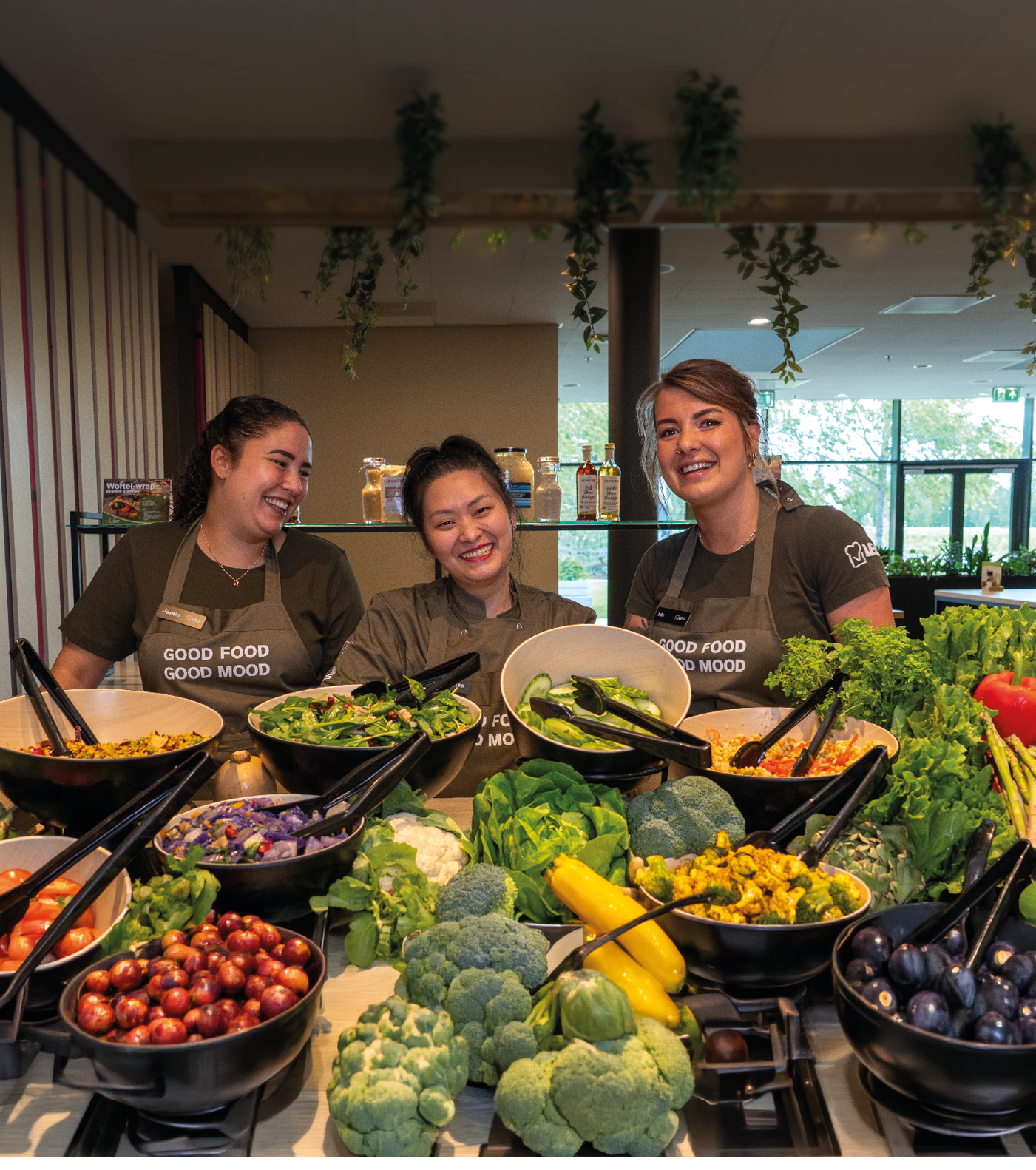 Three adults wearing matching aprons with the phrase “GOOD FOOD GOOD MOOD" smile behind a table filled with bowls of fresh vegetables and salad ingredients in a bright indoor setting. Description generated by AI