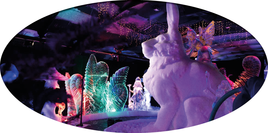 A colorful indoor ice sculpture display features a large rabbit with a fairy figure sitting on its back surrounded by illuminated abstract shapes and figures. Description generated by AI