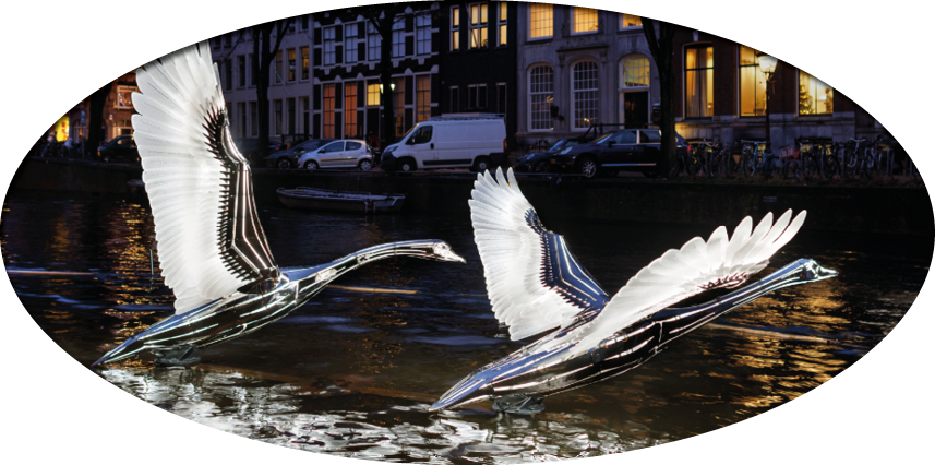 Two metallic bird sculptures appear to be flying over a canal with buildings and parked vehicles in the background at night. Description generated by AI