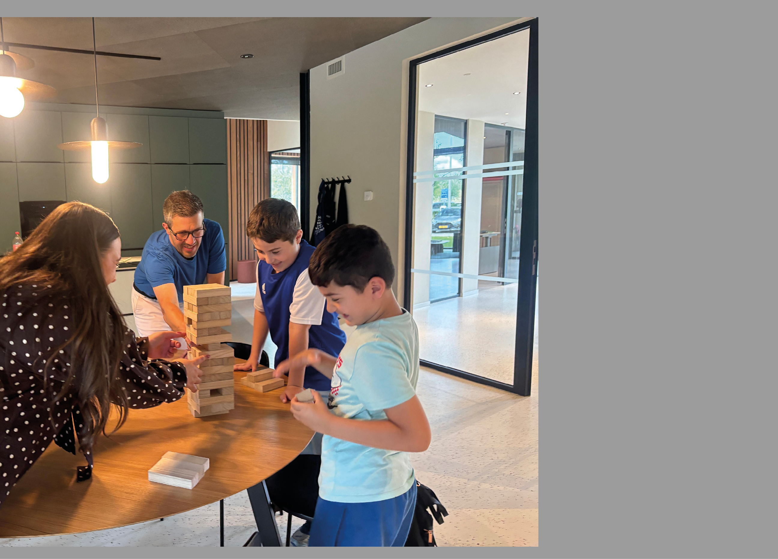 Four people are playing a game of Jenga around a wooden table in a modern indoor setting with large windows and pendant lights. Description generated by AI