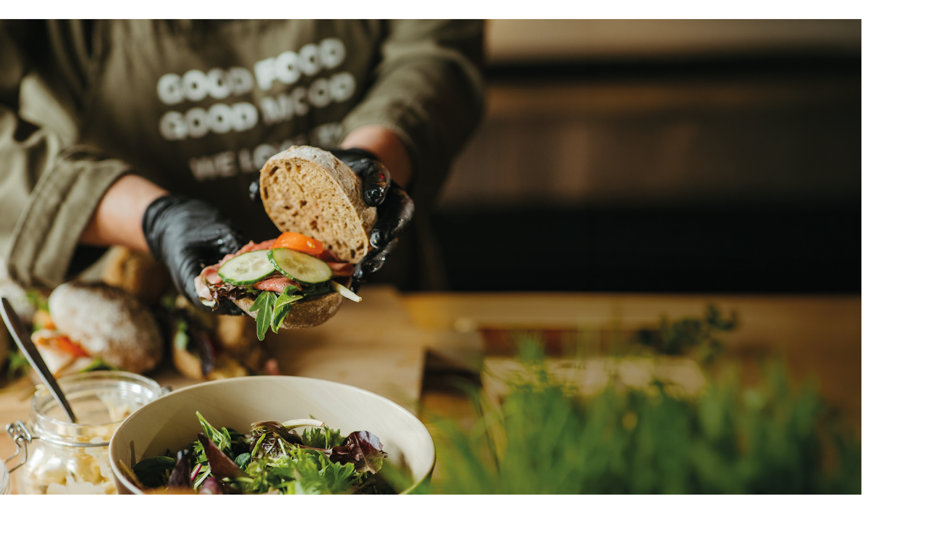 A person wearing black gloves is assembling a sandwich with fresh vegetables and greens over a wooden surface next to a bowl of salad. Description generated by AI