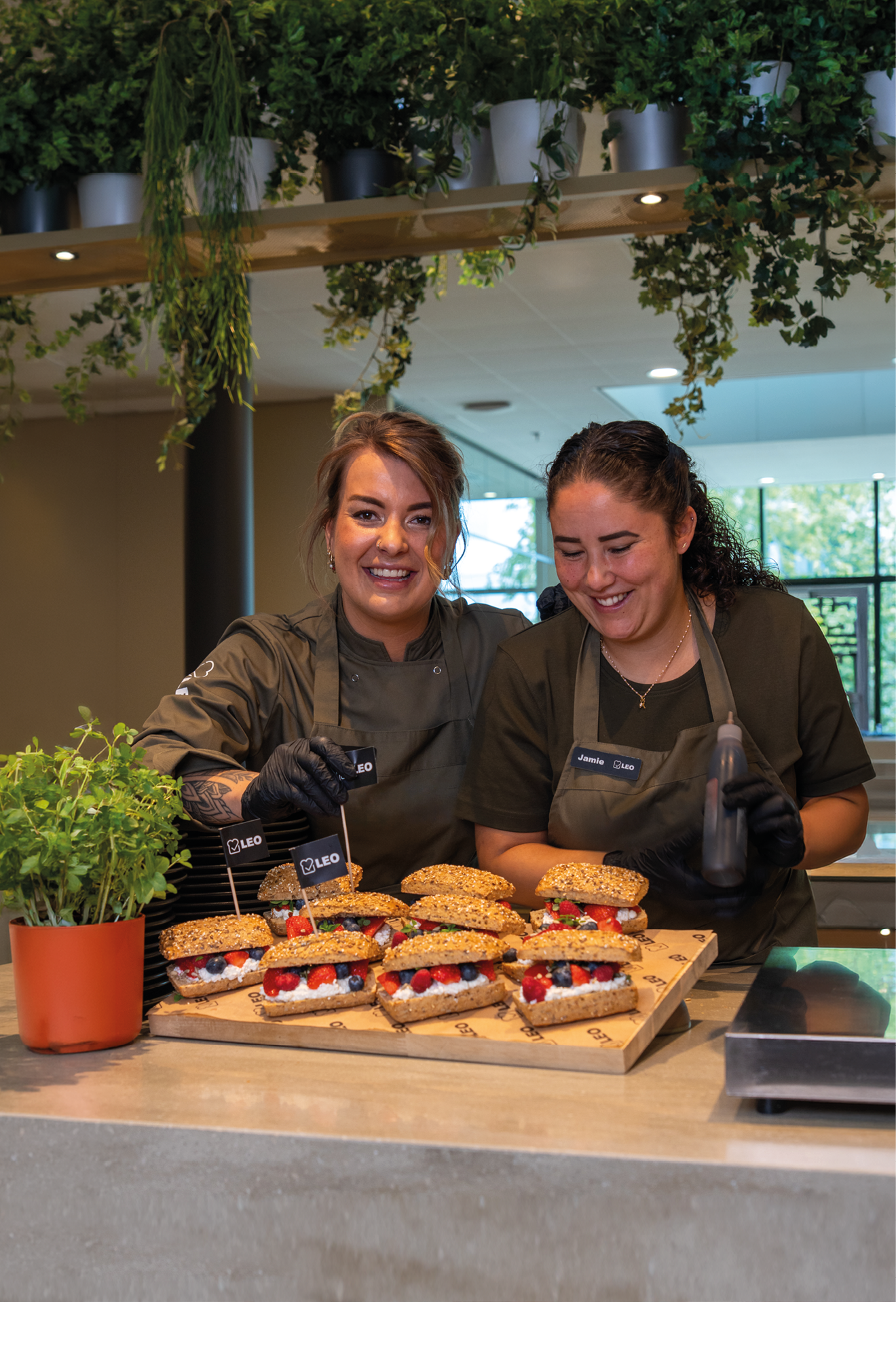 Two people wearing dark aprons and gloves smile while standing behind a counter displaying a wooden board with several sandwiches topped with small flags labeled “CIELO" in a bright, modern indoor setting with hanging plants. Description generated by AI