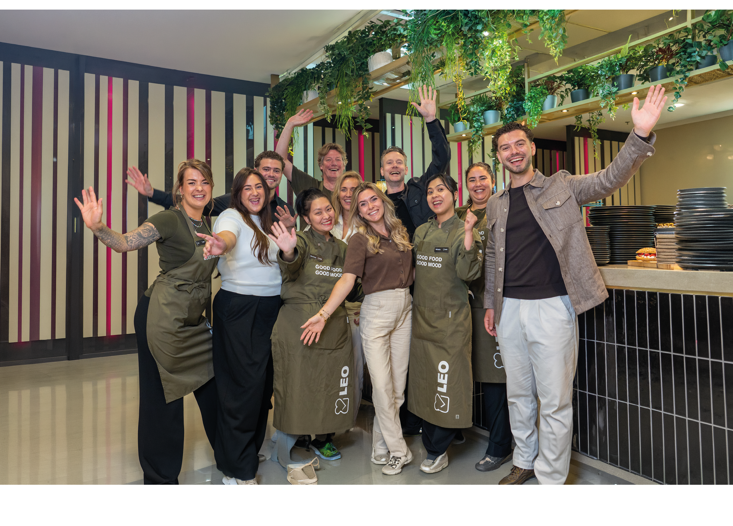 A group of smiling adults wearing aprons and casual clothing stand together indoors with their hands raised in a cheerful gesture under hanging plants. Description generated by AI