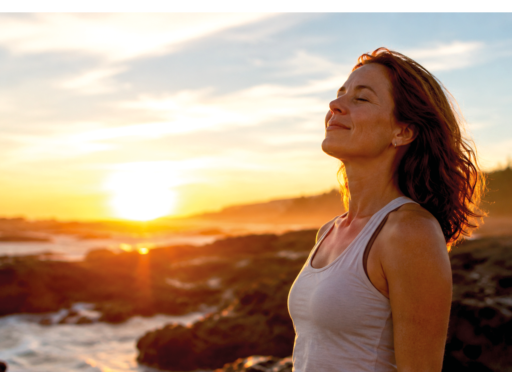 A woman with long hair and a white tank top standing on rocks near the ocean. Description generated by AI