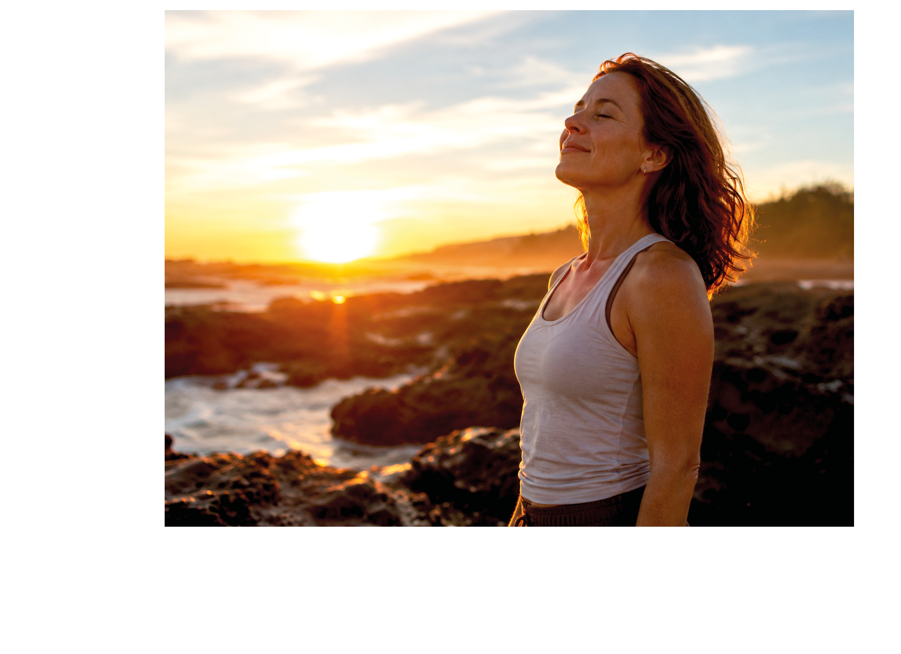 A woman with long hair and a white tank top standing on rocks near the ocean. Description generated by AI