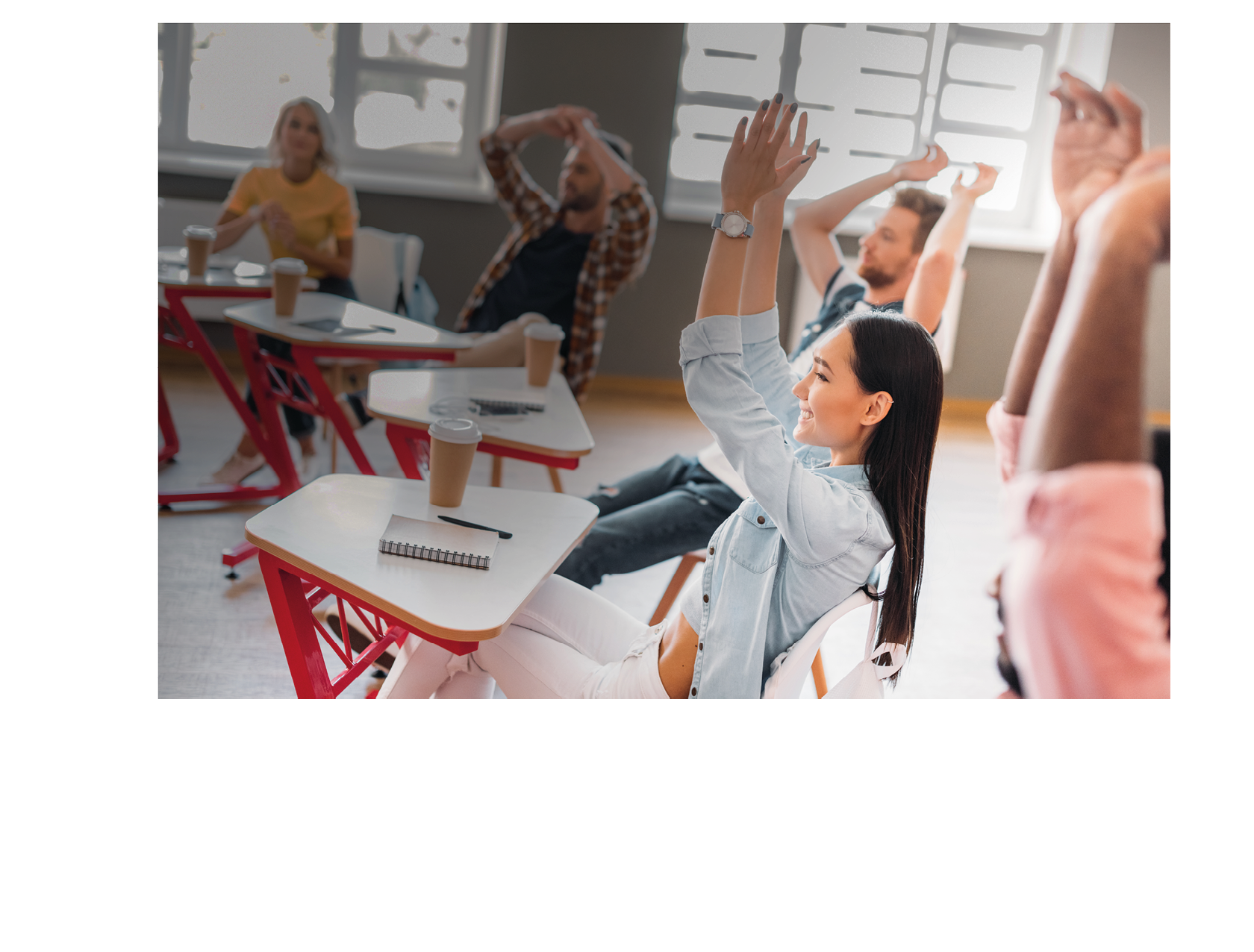 Adults sitting at desks in a classroom are stretching with their arms raised while smiling and engaging with each other. Description generated by AI