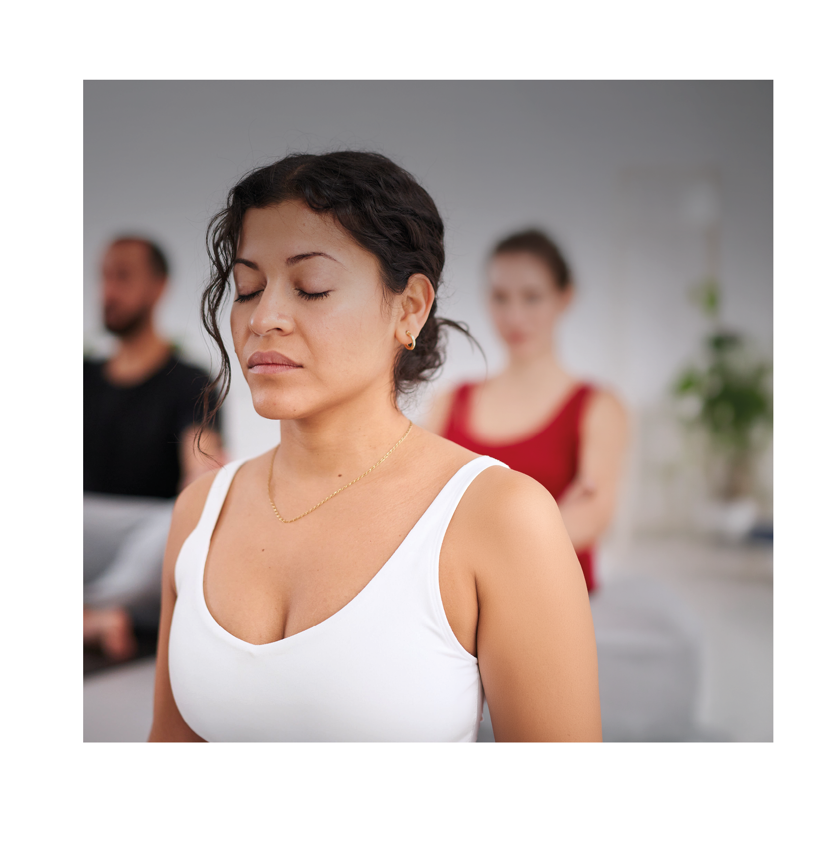 A person with curly hair wearing a white tank top is meditating with eyes closed while two other people sit quietly in the background. Description generated by AI